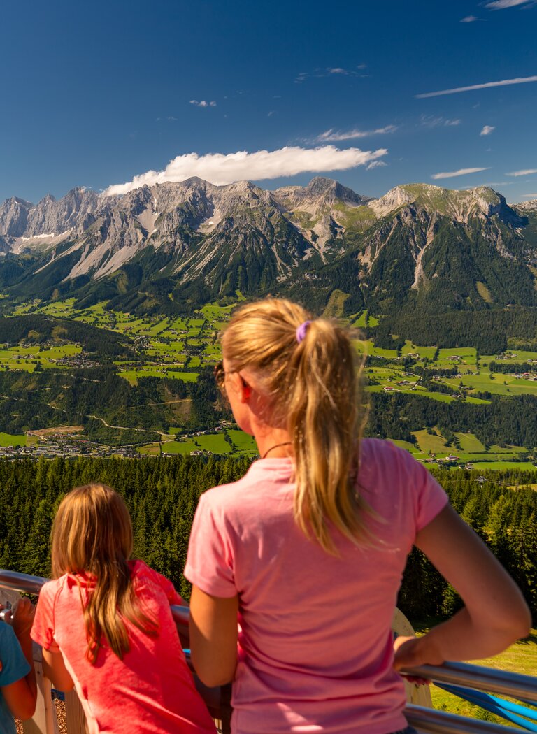 Family with backpack looks from viewing platform across sunny Enns Valley toward the impressive Dachstein mountain range | © Planai-Hochwurzen-Bahnen GmbH