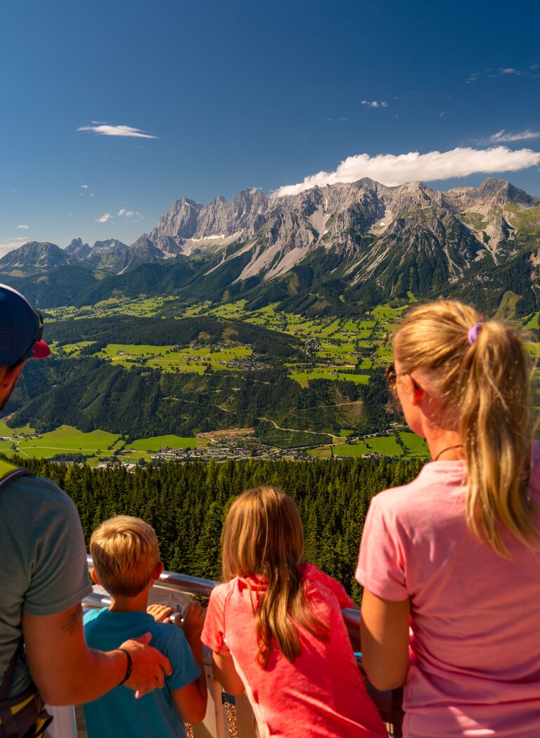 Family with backpack looks from viewing platform across sunny Enns Valley toward the impressive Dachstein mountain range | © Planai-Hochwurzen-Bahnen GesmbH