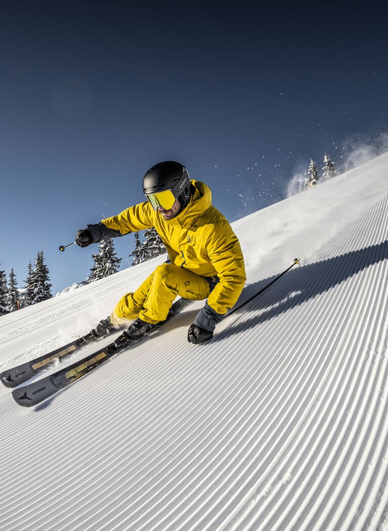 Athletic skier in yellow outfit carves down a freshly groomed slope under bright sun in a snowy mountain ski resort | © Planai-Hochwurzen-Bahnen GmbH
