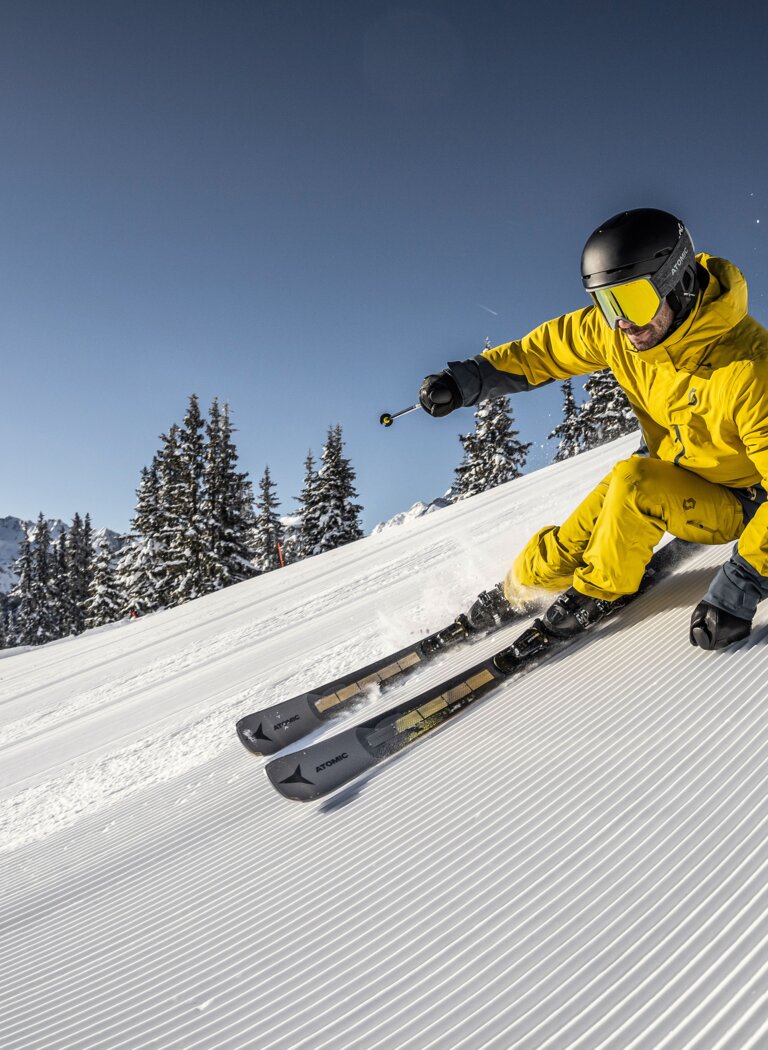 Athletic skier in yellow outfit carves down a freshly groomed slope under bright sun in a snowy mountain ski resort | © Planai-Hochwurzen-Bahnen Gesellschaft m.b.H.