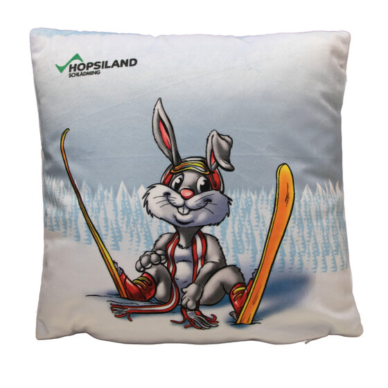 Square pillow with bunny Hopsi in ski gear in front of snowy forest, yellow skis and 'Hopsiland' print | © Planai-Hochwurzen-Bahnen GmbH