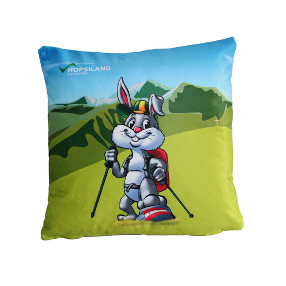 Quadratisches Kissen mit bunter Berglandschaft und Wanderhase Hopsi mit Rucksack und Stöcken, Aufdruck 'Hopsiland' | © Planai-Hochwurzen-Bahnen GmbH