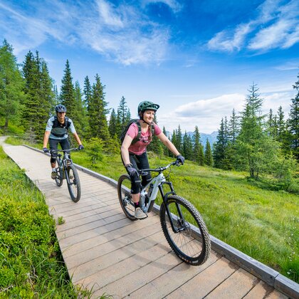 Two mountain bikers ride a wooden path through flowery alpine meadows and forest, wearing backpacks and protective gear | © Josh Absenger