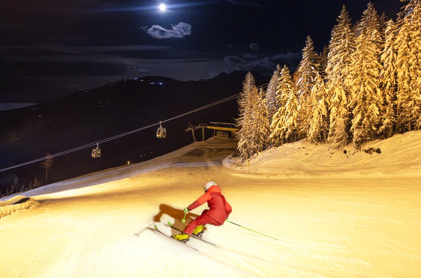Nachtskifahren unter dem Vollmond mit beleuchteter Piste | © Planai-Hochwurzen-Bahnen Gesellschaft m.b.H. Skifahrer in rotem Anzug fährt nachts auf beleuchteter Piste, umgeben von verschneiten Bäumen und Gondelbahn bei Vollmond. | © Planai-Hochwurzen-Bahnen Gesellschaft m.b.H.