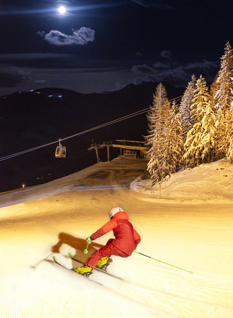 Skier in red suit descends illuminated slope at night, surrounded by snowy trees and gondolas under a bright full moon. | © Martin Huber