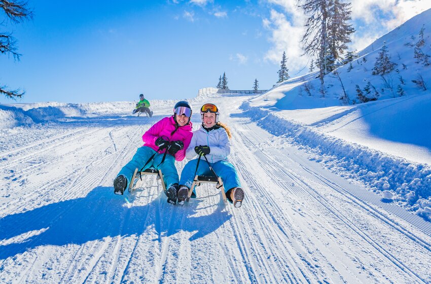 Zwei lachende Mädchen rodeln nebeneinander eine frisch präparierte Schneepiste hinunter, bei Sonne und Pulverschnee | © Planai-Hochwurzen-Bahnen Gesellschaft m.b.H.