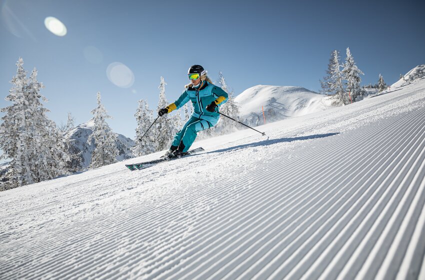 Skier in turquoise outfit skiing on a freshly groomed slope under blue sky and bright sunshine. | © Reiteralm Bergbahnen GmbH & Co.KG