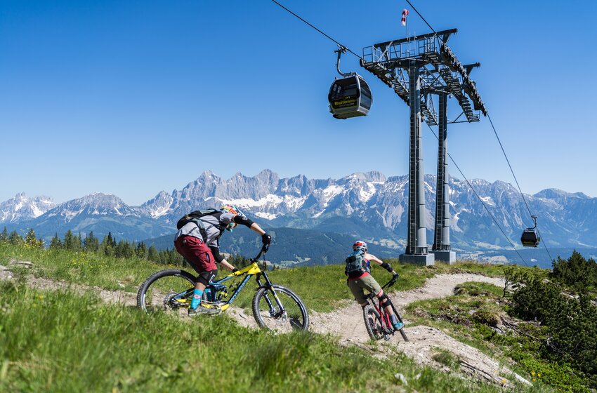 Zwei Mountainbiker fahren einen kurvigen Trail unter einer Gondelbahn vor schneebedeckten Alpen und blauem Himmel. | © Reiteralm Bergbahnen GmbH & Co.KG