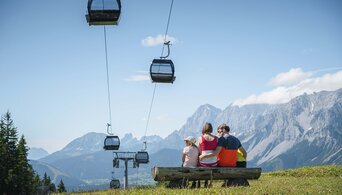 Family sitting on a bench overlooking gondolas and the impressive Dachstein mountain range. | © Ski amadé GmbH
