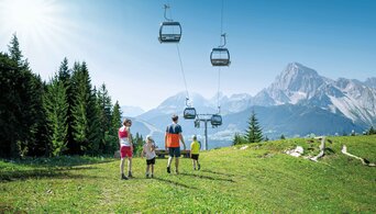 Family hiking through green alpine meadow with gondola and mountain backdrop on a sunny day. | © Ski amadé GmbH