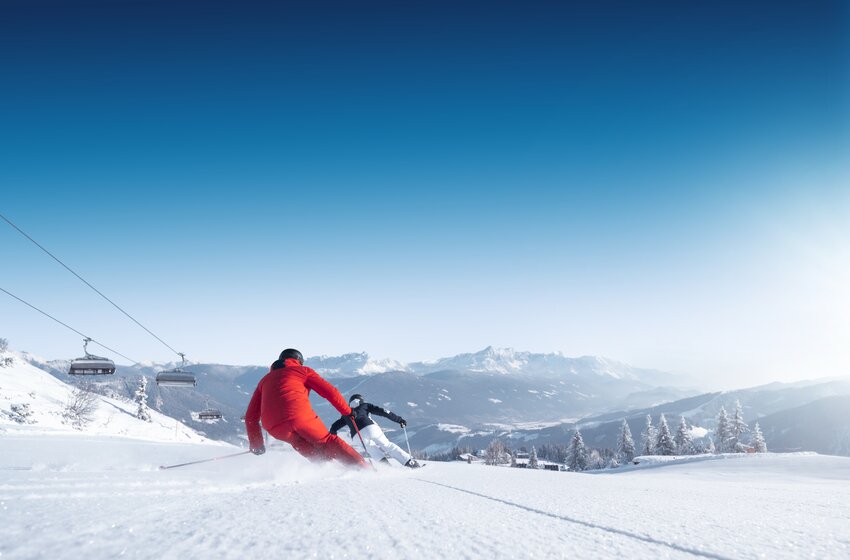 Two skiers in red and black carve on an empty slope with a wide view over the snowy valley and mountains in Ski amadé. | © Ski amadé GmbH
