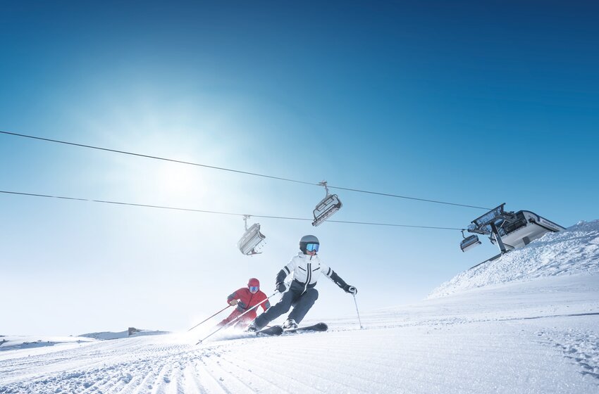 Zwei Skifahrer carven auf frisch präparierter Piste unter Sessellift bei blauem Himmel und strahlender Morgensonne. | © Ski amadé GmbH